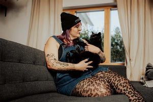 Leo-Strumpfhose an den Beinen einer Frau, die eine Mütze und ein Jeanskleid trägt. Sie hat viele Tattoos und hält einen schwarzen Kater im Arm.