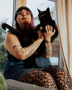 Strumpfhose mit Leomuster an Plus Size Model mit pinken Haaren, Mütze, Jeanskleid und Tattoos. Sie hält einen schwarzen Kater auf dem Arm.