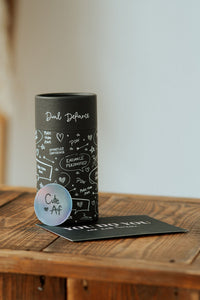 Dual Defiance PaperTube Verpackung für Strumpfhosen und Strümpfe. Trendig, wiederverwendbar oder als Geschenk. eine schwarze Papierdose steht auf dunklem Holz, davor ein runder Holo-Sticker und eine schwarze Postkarte.