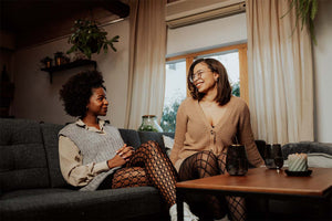 Zwei Frauen in eleganten schwarzen Strumpfhosen, Netzstrumpfhose und Feinstrumpfhose. Sie schauen sich lachend an und sitzen auf einem Sofa.