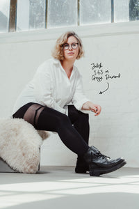 Die Frau sitzt auf einer mit Kunstfell bedeckten Bank mit einem ausgestreckten Bein. Sie trägt ein weißes Hemd, eine große Brille mit runden Gläsern, lockere schwarze Boots und Strumpfhosen.